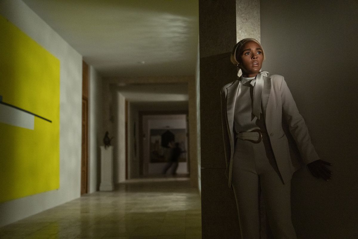 Janelle Monáe looks scared in a dark empty corridor with modern art in the wall. In the background a shadowy figure can just be seen