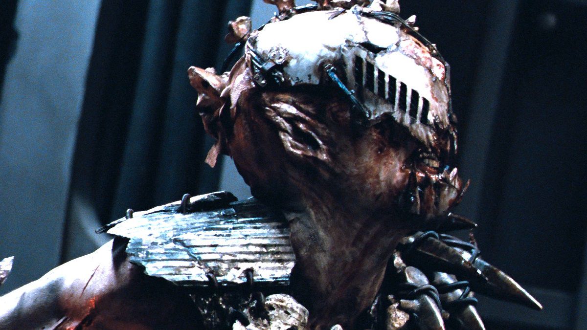 Un humano mutante macabro con rasgos faciales visiblemente mutilados y púas de metal que sobresalen de su carne enseñando los dientes a algo fuera de marco.