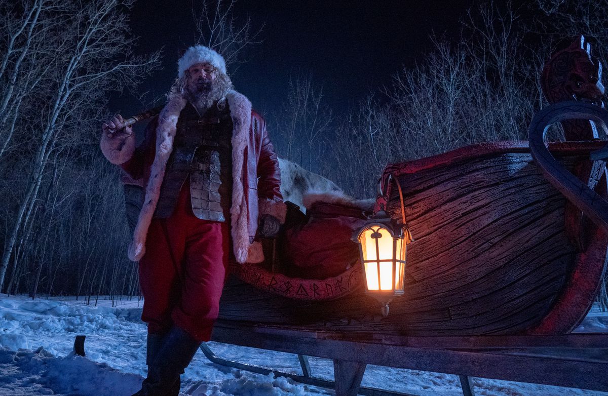 Санта-Клаус (Дэвид Харбор) пьяно прислоняется к своим саням, кроваво-красному деревянному транспортному средству в форме лодки, украшенному скандинавскими рунами, в «Жестокой ночи».