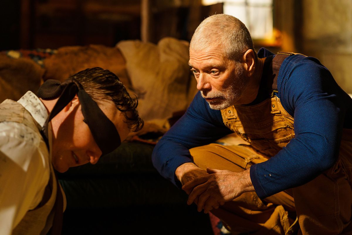 Um velho (Stephen Lang) em uma camisa azul de manga comprida e macacão amarelo agacha-se na frente de um homem vendado (Marc Senter).