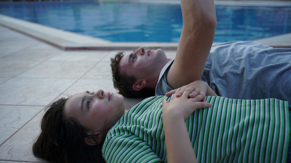 Een vader en dochter liggen op hun rug naast een zwembad terwijl de vader in Aftersun naar de lucht wijst.