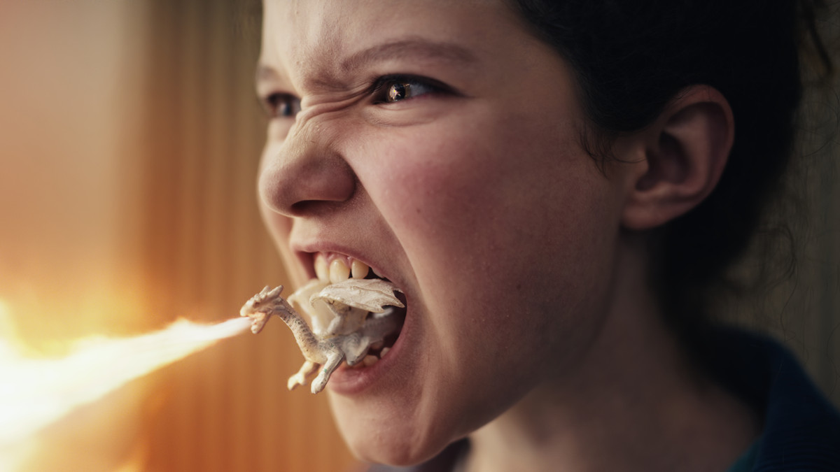 Ein winziger weißer Drache schießt eine Feuerfahne aus seinem Mund, während er im offenen Mund eines sichtlich wütenden jungen Mädchens sitzt.