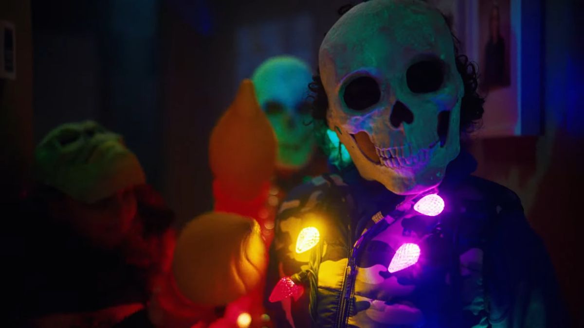 Uma pessoa usando uma máscara com cara de caveira e um traje elaborado com luzes multicoloridas espalhadas em volta do pescoço.