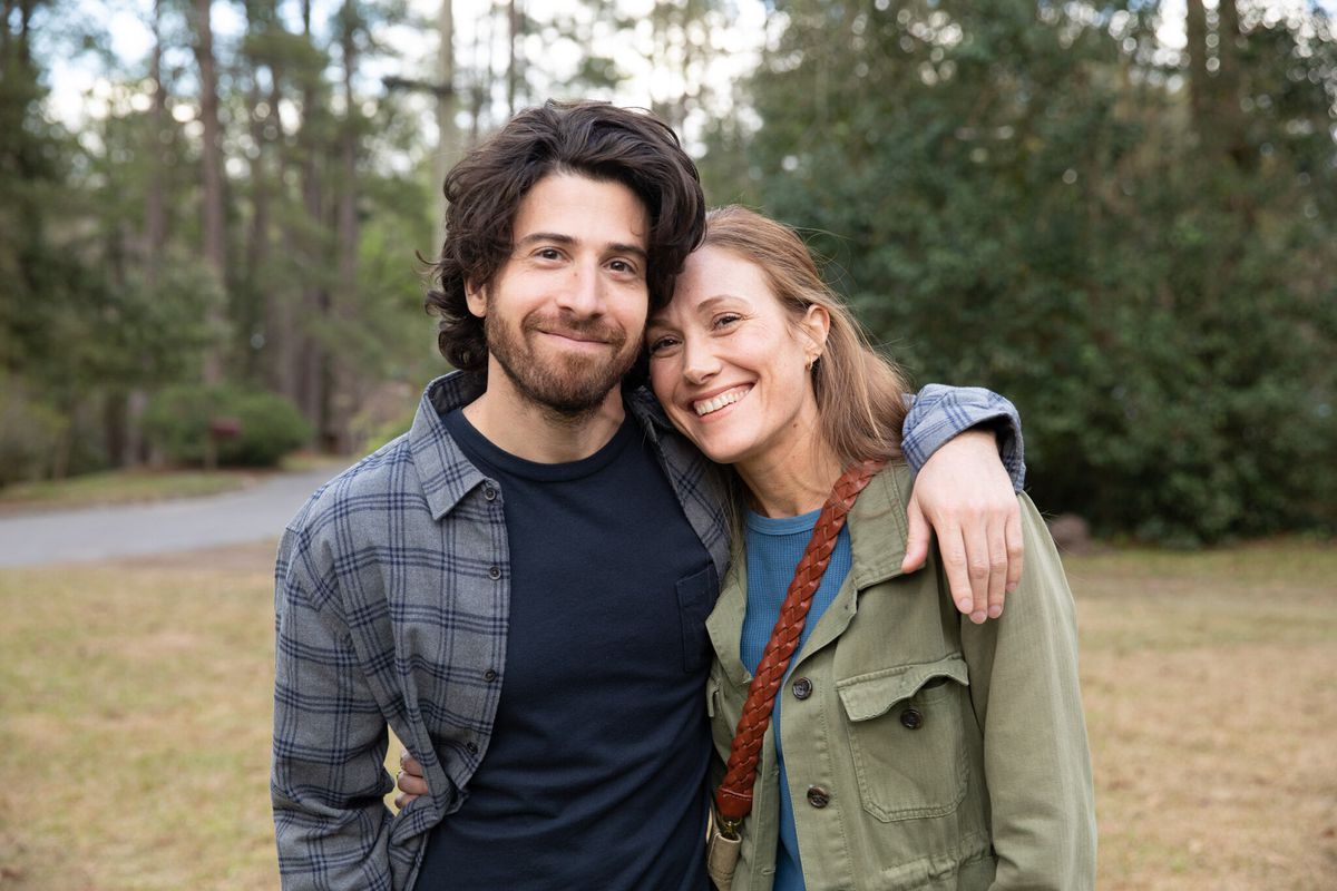 Um homem (Jake Hoffman) em uma camisa azul escura posa para uma foto com uma mulher sorridente (Schuyler Fisk) em uma jaqueta verde oliva.