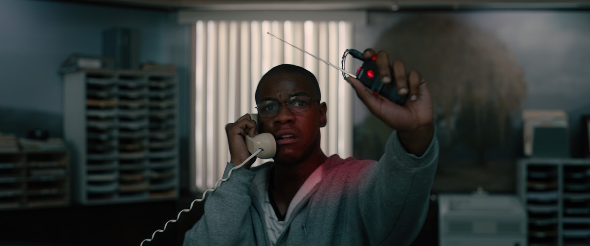 John Boyega usa una sudadera con capucha gris y sostiene un detonador en una mano mientras habla por teléfono con la otra en Breaking.