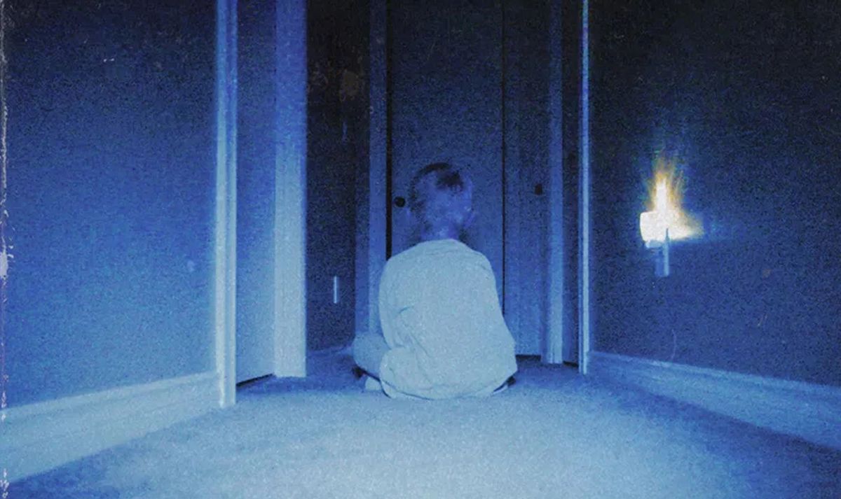 一个小男孩背对着镜头坐在昏暗的蓝色走廊里，面对一系列敞开的门口，这是恐怖电影 Skinamarink 中典型的模糊模糊镜头