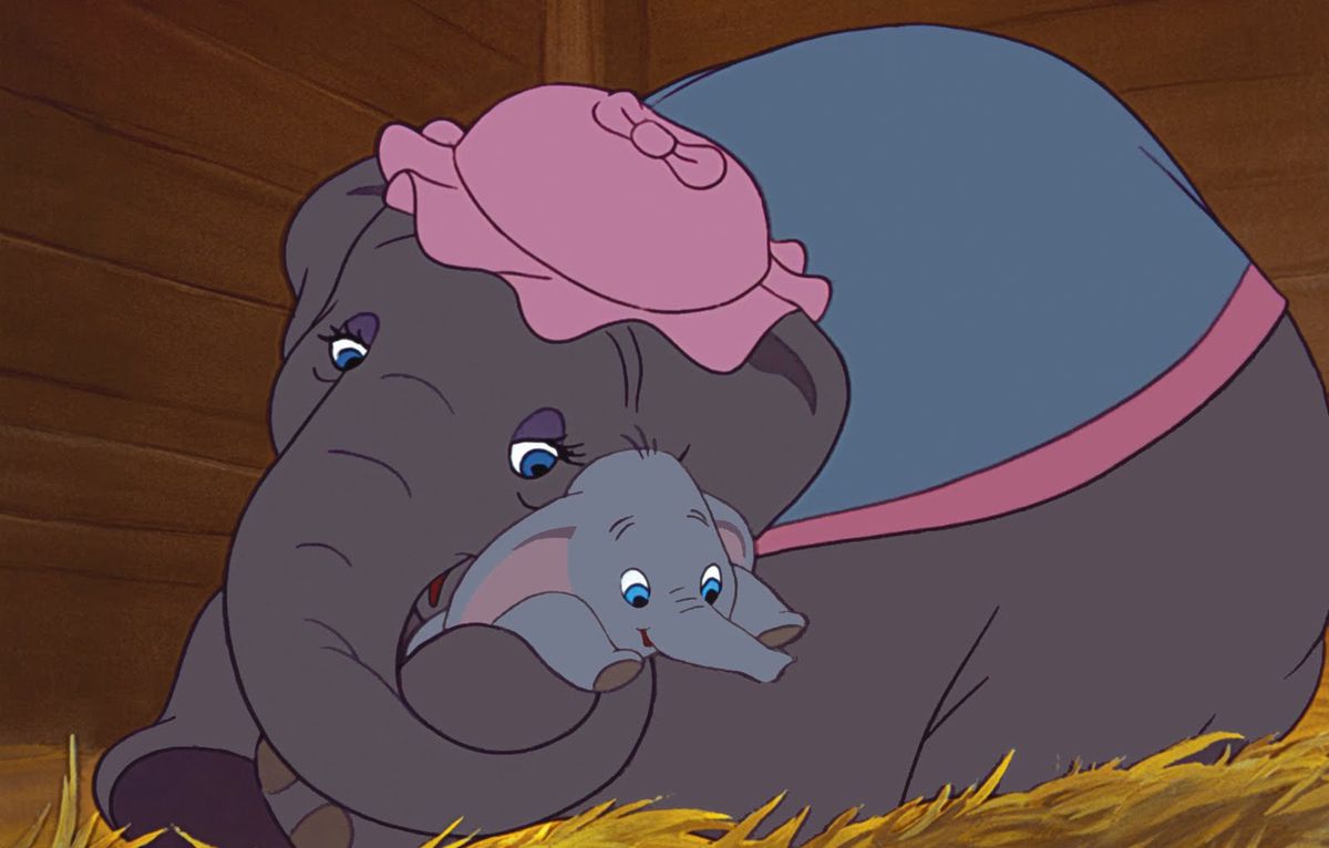 Mrs. Jumbo, an elephant in a pink mob cap and blue shawl, cradles her baby Dumbo in her trunk while lying in a bed of straw in 1941’s animated feature Dumbo
