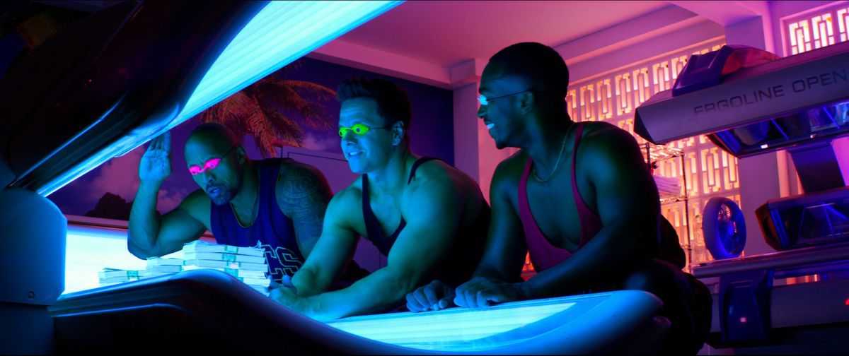 在《痛苦与收获》中，道恩·强森、马克·沃尔伯格和安东尼·麦基都穿着五颜六色的背心在晒黑床前微笑。