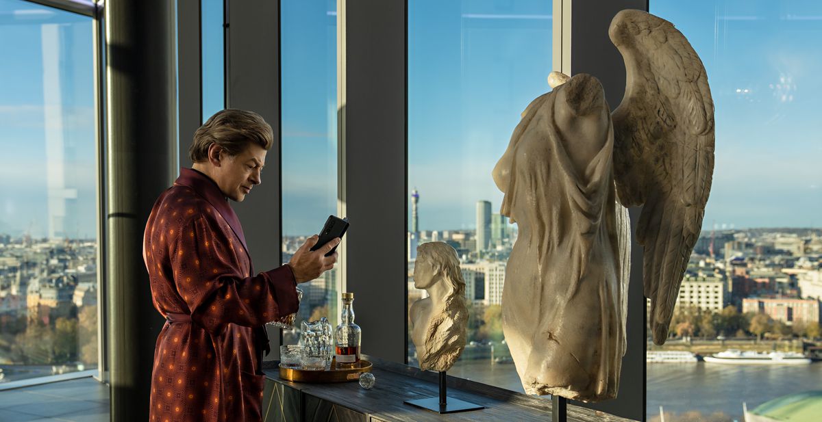 Billionaire criminal David Robey (Andy Serkis, in burgundy bathrobe and a thick, wavy wig) stands by a wall of windows overlooking London, with a headless, armless angel statue in the foreground, in Luther: Fallen Sun