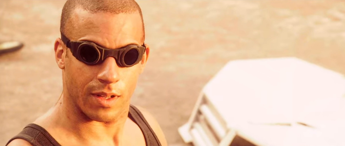 Vin Diesel as Richard B. Riddick facing the camera wearing goggles and a black tank top with sand in the background