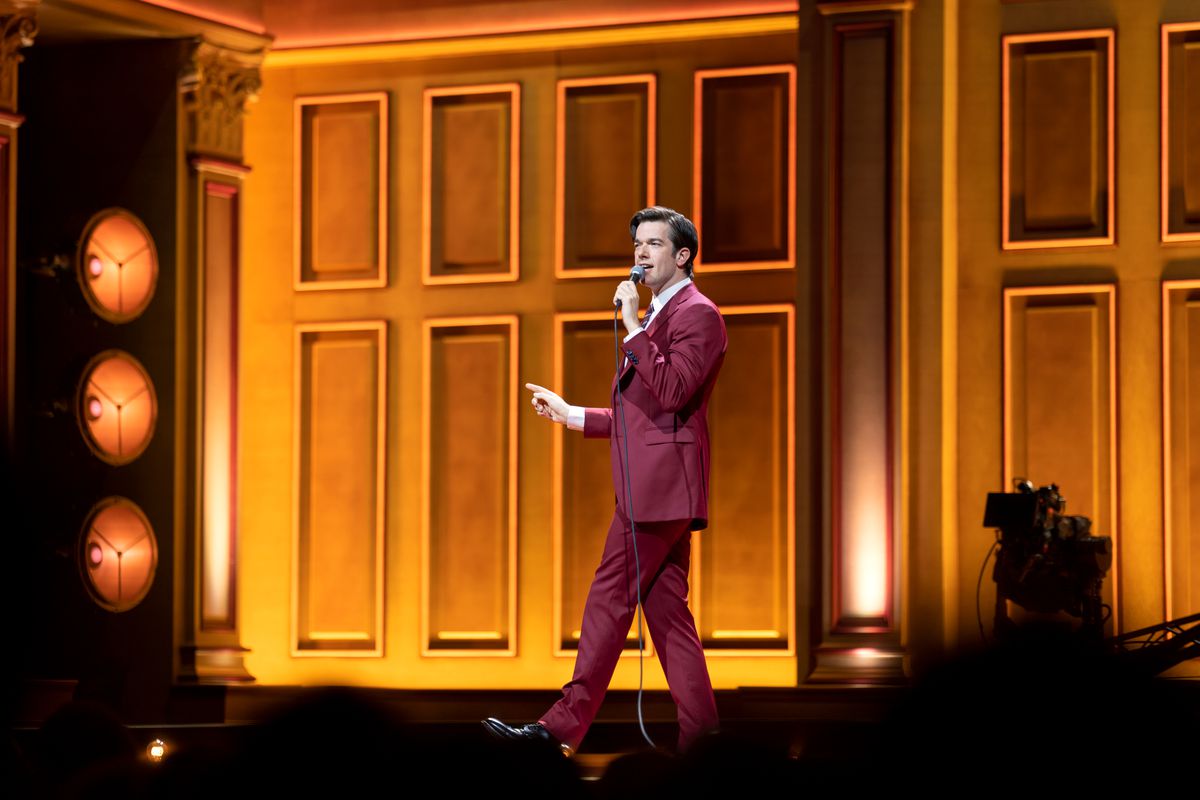A wide shot of comedian John Mulaney as he walks stage right against an orange background in a red suit in his Netflix special Baby J.