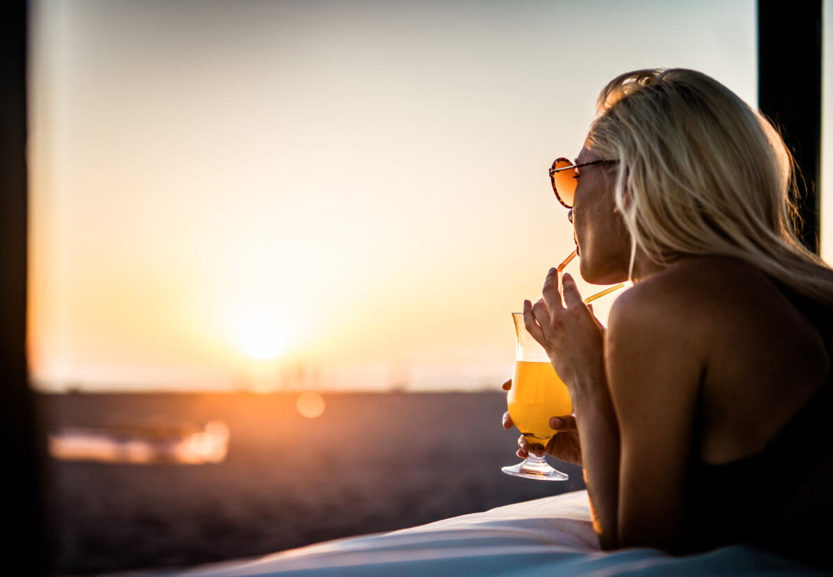 Ung kvinne drikker sommercocktail på strandsengen ved solnedgang.