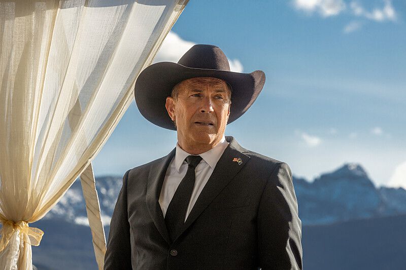 Kevin Costner in Yellowstone, standing facing the camera wearing a black suit with a black cowboy hat