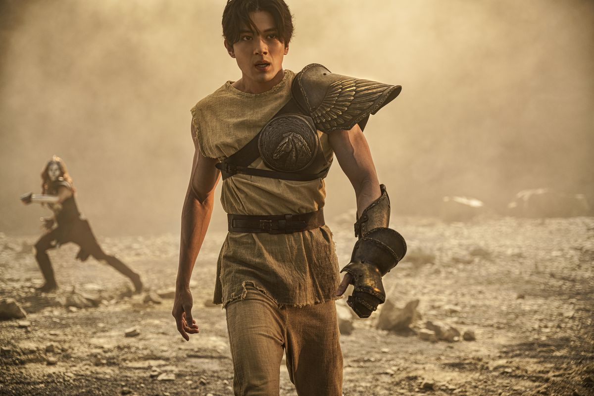 Seiya, the protagonist of the live-action 2023 Knights of the Zodiac, walks across a sandy, windblown space while wearing a brown tunic, a single metal gauntlet, and a wing-shaped brass pauldron