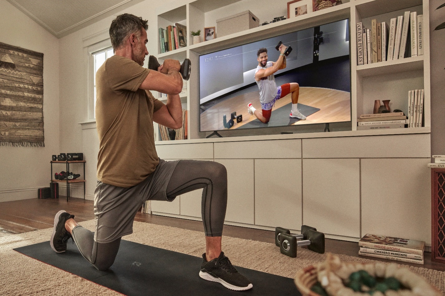 Man working out with a weight while viewing a Peloton class on his TV in a living room.