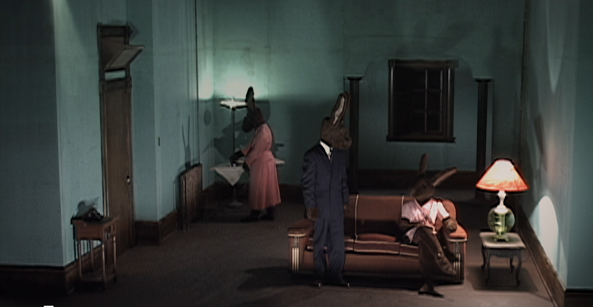 Several human sized rabbits dressed in human clothes stand in a living room in David Lynch’s Inland Empire