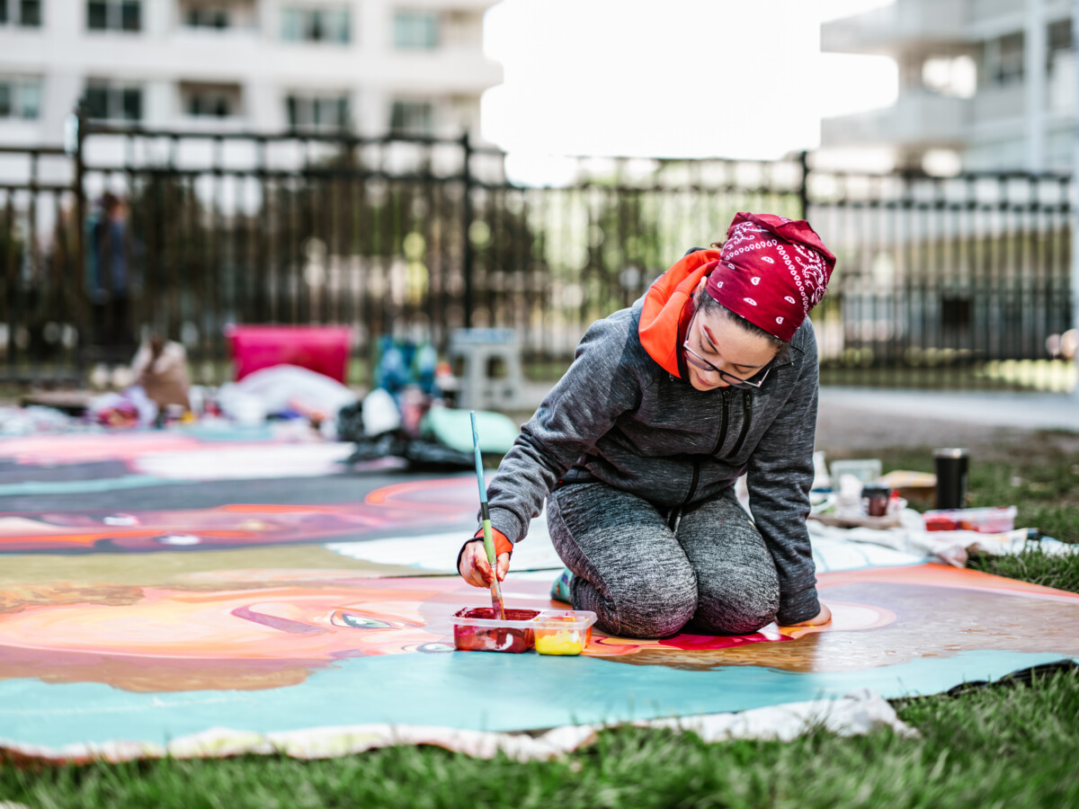 mujer creando un mural al aire libre en el césped. Está vestida con ropa informal, con pañuelo y anteojos. Exterior del parque público de la ciudad.