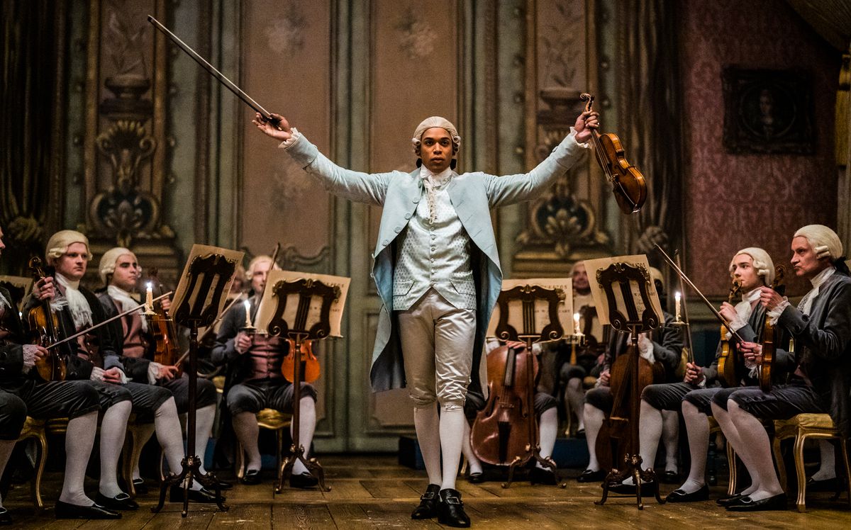 Kelvin Harrison Jr. holds up his violin while standing in front of an orchestra in Chevalier