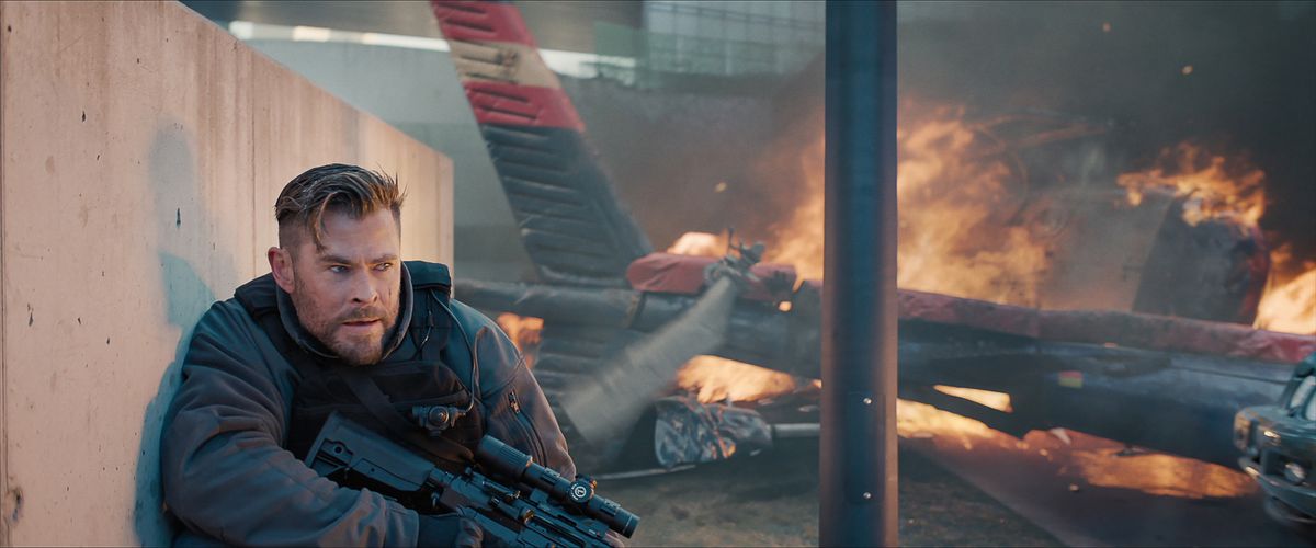 Mercenary Tyler Rake (Chris Hemsworth) crouches against a concrete wall and holds onto a heavy sniper rifle as the wreckage of a helicopter burns beside him in Extraction 2