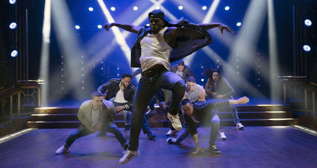 Magic Mike's Last Dance의 한 장면에서 밝은 흰색 스포트라이트가 교차하는 파란색 조명 무대에서 다른 남성 댄서들이 그의 뒤에 웅크리고 있을 때 청바지와 뒤로 야구 모자를 쓴 흑인 남성 댄서가 공중 높이 뛰어오릅니다.