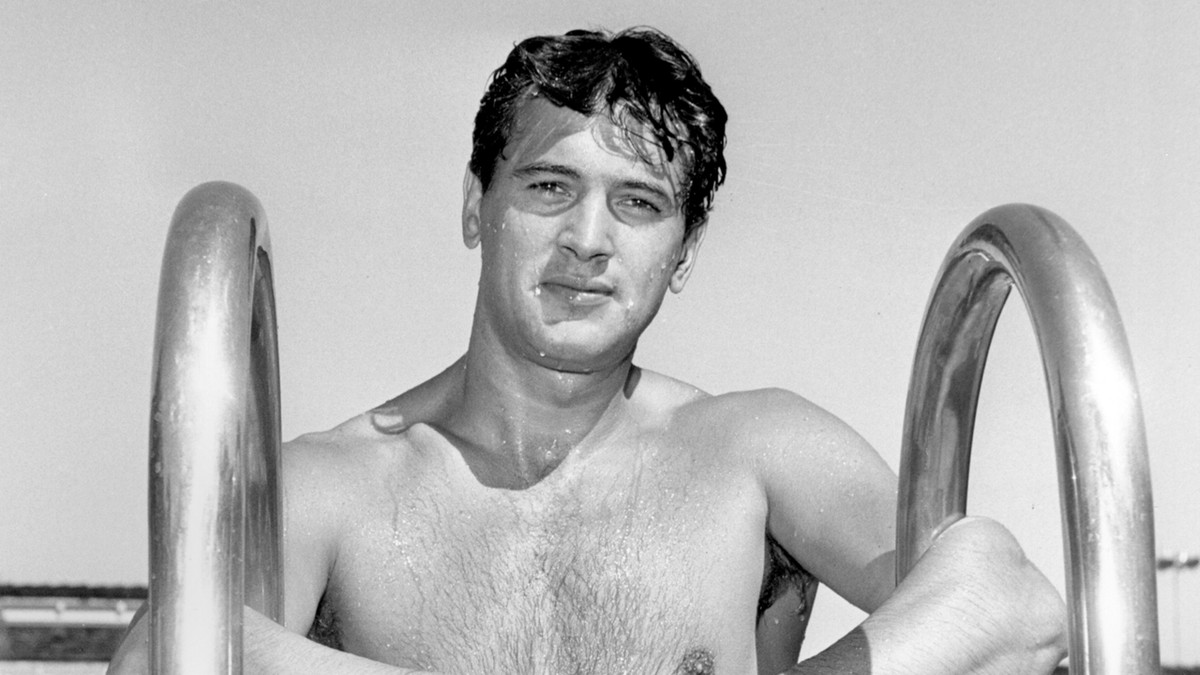 A black-and-white archival photo of Rock Hudson at the top of a pool ladder.