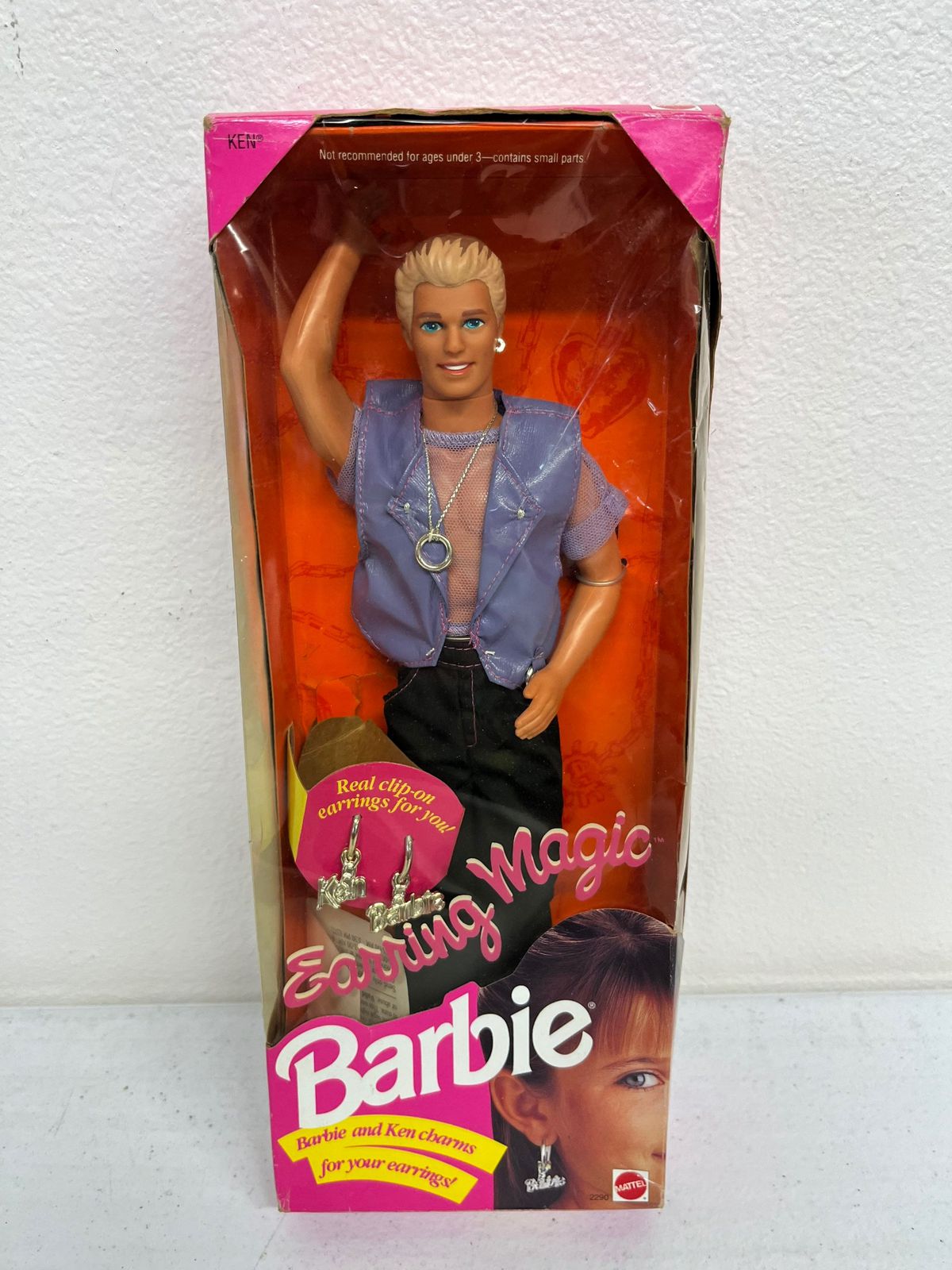 A product shot of Earring Magic Ken in the box: A Ken doll with brown-and-blonde streaked hair, a purple mesh shirt and purple pleather vest, and a very visible silver ring on a chain around his neck