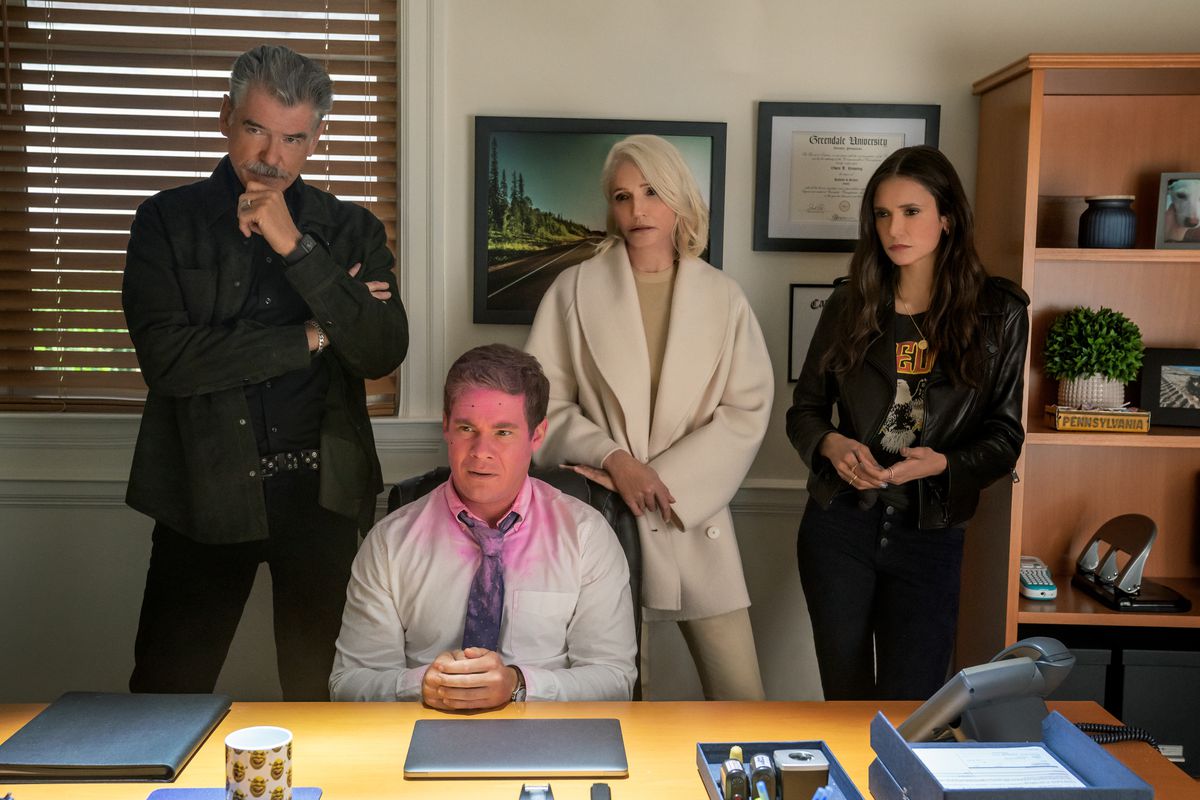 A young man sits at a desk, hands folded, pink paint all over his face. Behind him are a cool older couple — a silver-haired man in leather, and a sleek lady in white — and a young woman in a cool leather jacket. 
