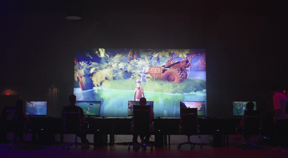 A group of people sit at computer stations in a dark room in front of a giant screen with a game scene display on it.