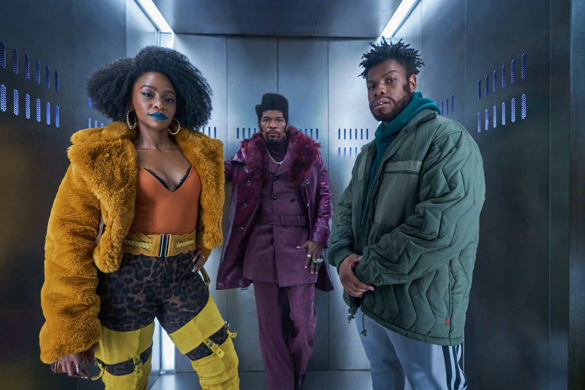 (L-R) Teyonah Parris in a orange fur coat, orange body suit with leopard print paints, Jamie Foxx in an all-purple suit with matching coat, and John Boyega in a puffy teal coat standing in a metallic elevator in They Cloned Tyrone.