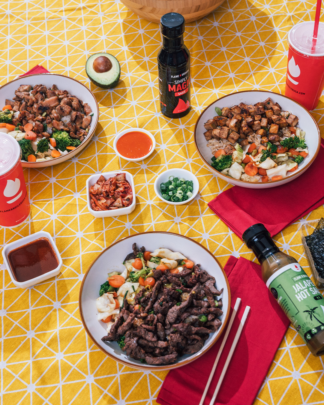 rice bowls with meat and vegetables, scallions, kimchi, chopsticks, sauces and drinks on a table with a yellow tablecloth at a the flame broiler fundraiser