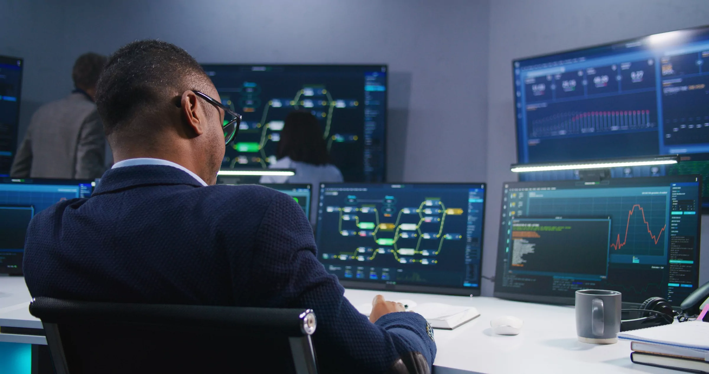 Software programmer sits in front of computer with blockchain network and data server in monitoring office. Team of IT technical specialists work with live analysis charts on big digital screens.