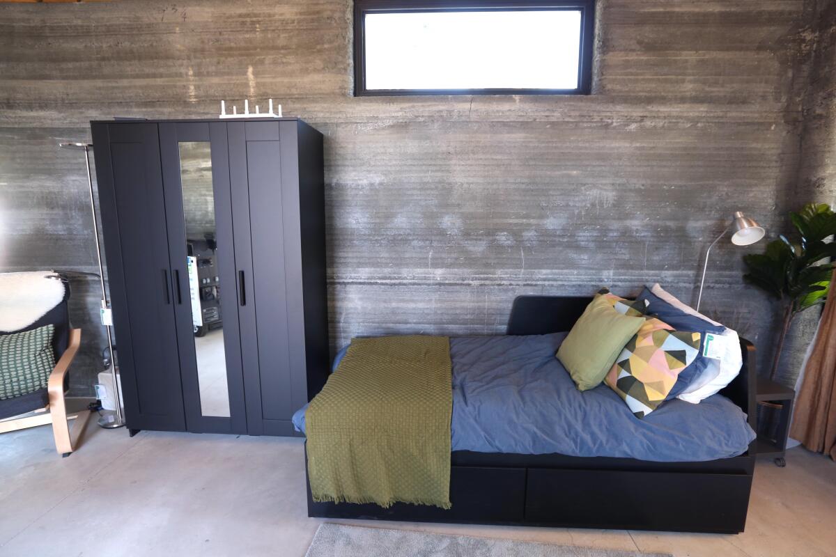A bed, armoire and chair are shown before a 3-D-printed concrete wall that has an almost geological texture.