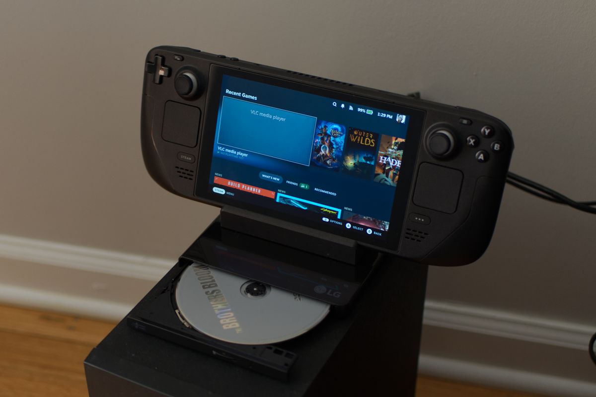 An image showing the Steam Deck portable console nested into its TV dock. The Deck and its dock are sitting on top of an external DVD player that is plugged into the dock.