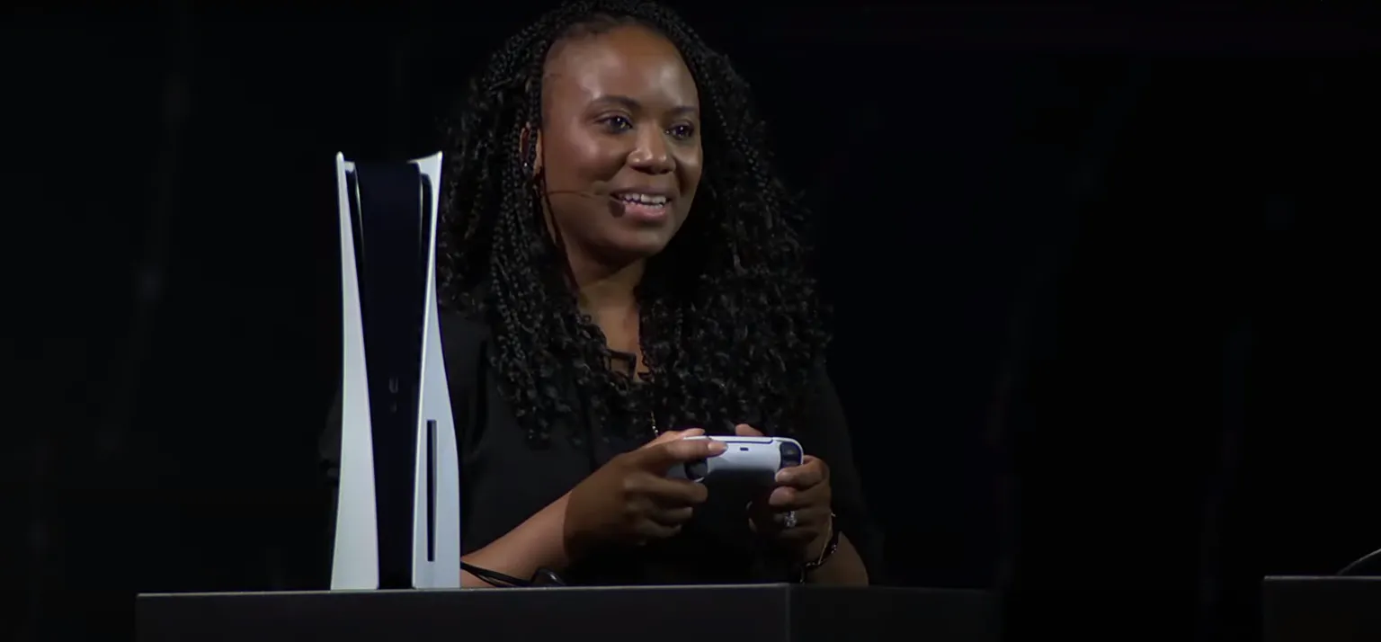 لقطة شاشة تظهر امرأة تحمل وحدة تحكم PS5 ووحدة تحكم PS5 بجانبها. خلفية سوداء.