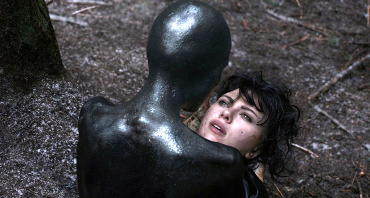 A black humanoid creature holds a human face in their hands in Under the Skin.