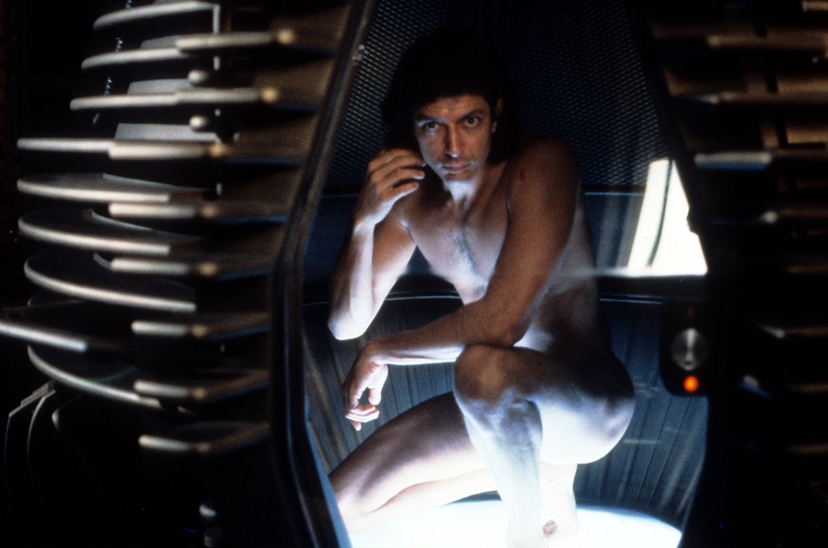 Jeff Goldblum, gol in ostro osvetljen, čepi v zloveščem, futuristično videti kovinskem podu v prizoru iz filma The Fly iz leta 1986