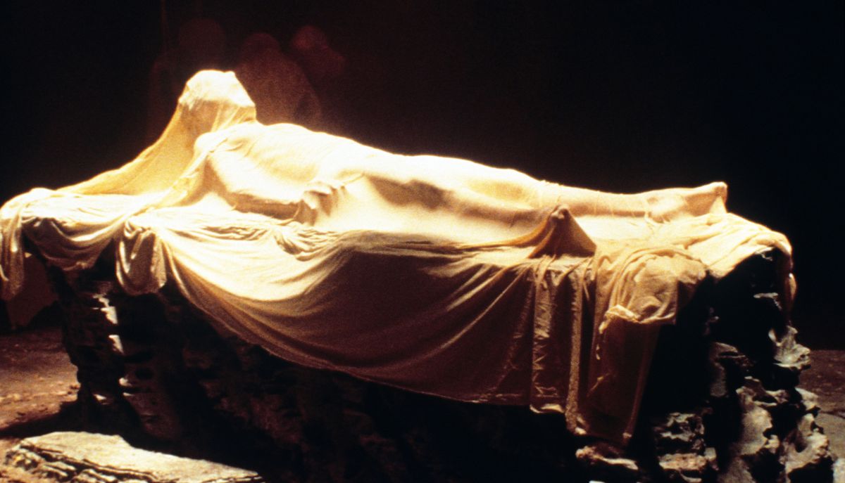 A human figure lies on a table and screams, completely blanketed in a yellow-white sheet-like cover that clings closely to his chest, legs, and open mouth in Fire in the Sky