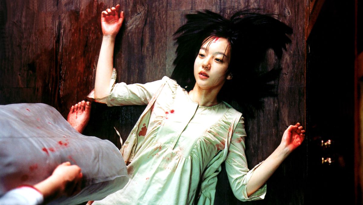 En koreansk kvinna med blodiga händer och fläckar av blod på sin klänning ligger på rygg på golvet, håret spritt runt henne i en gloria, som en figur med bara, blodiga fötter står över henne i A Tale of Two Sisters
