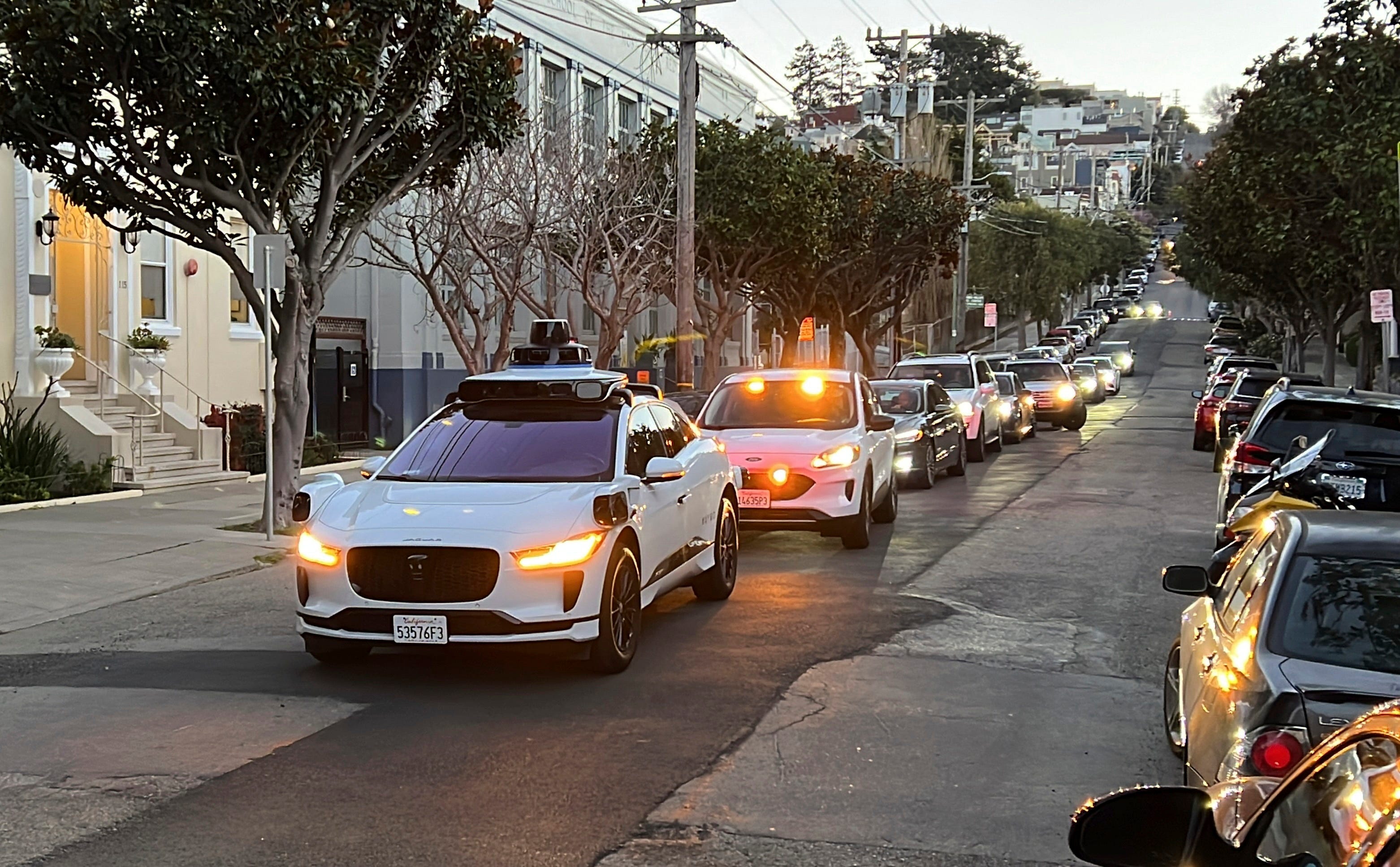 een witte auto blokkeert een rij auto's die erachter wachten in een stadsstraat. Het is een zelfrijdende Waymo-taxi.