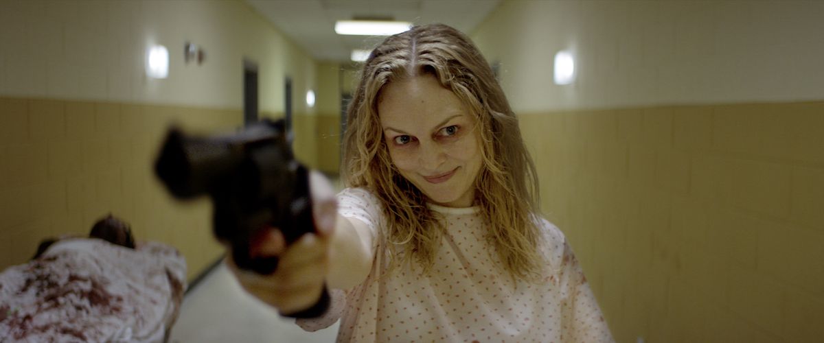 Elizabeth (Heather Graham) points a gun into the camera with a terrible smirk as she stands in a yellowing hospital hallway in a hospital robe in Suitable Flesh