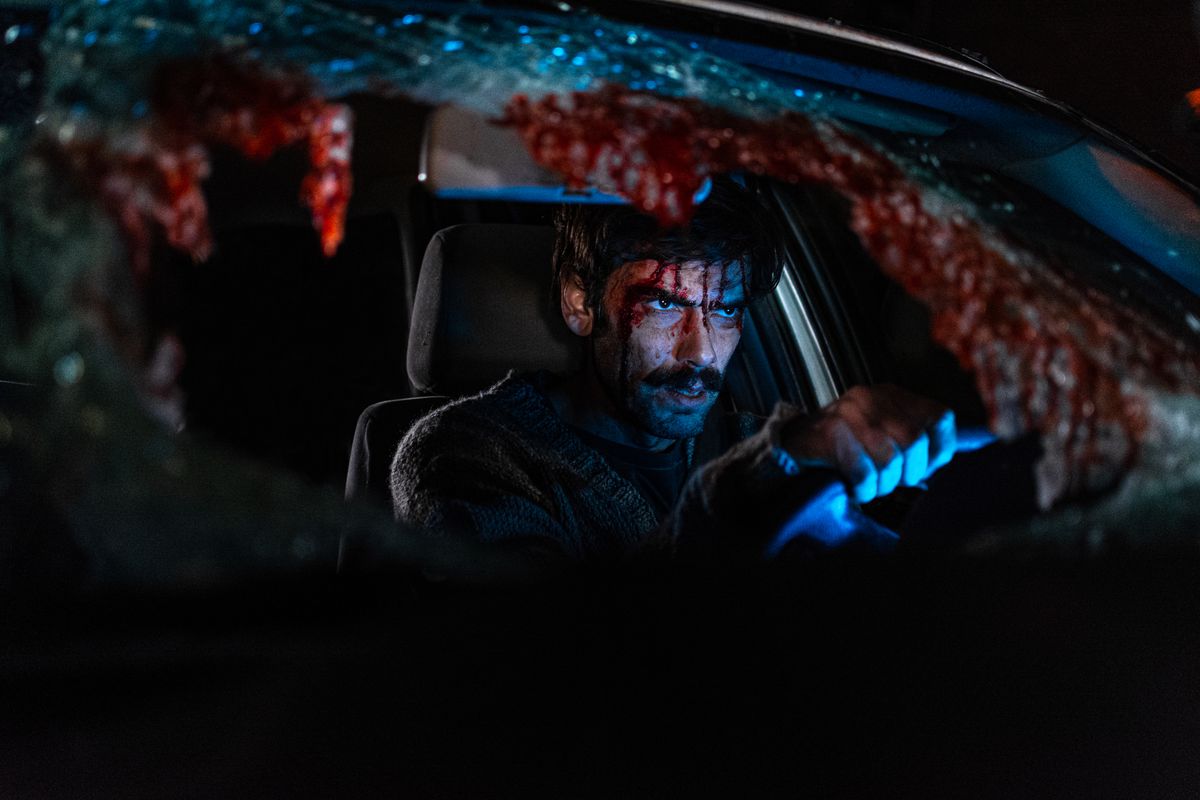 Ezequiel Rodríguez, with his face covered in blood, sits in the driver’s seat of a car with his hands on the wheel in When Evil Lurks. The car’s front windshield is shattered, with lots of blood.