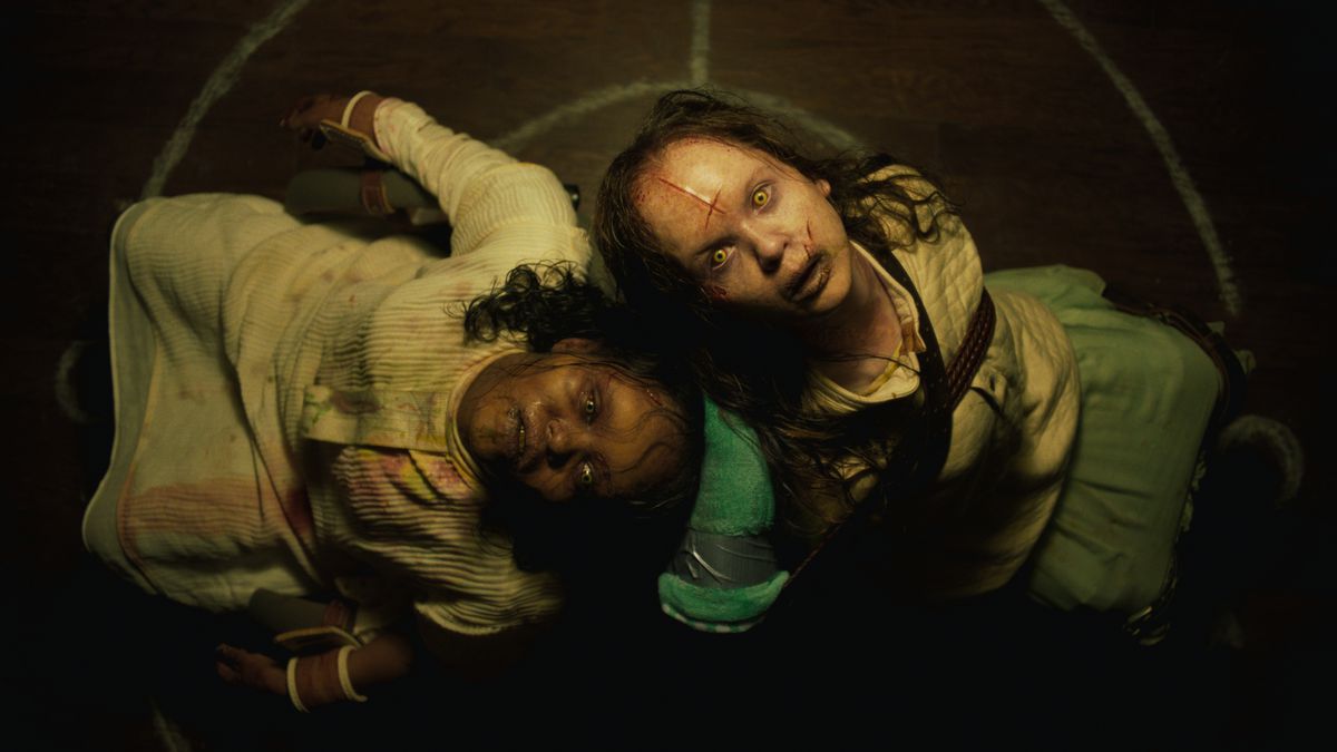 دو کودک تسخیر شده، زخمی و کبود شده پشت هم روی زمین نشسته اند و به دوربین بالای سرشان در The Exorcist: Believer خیره می شوند.