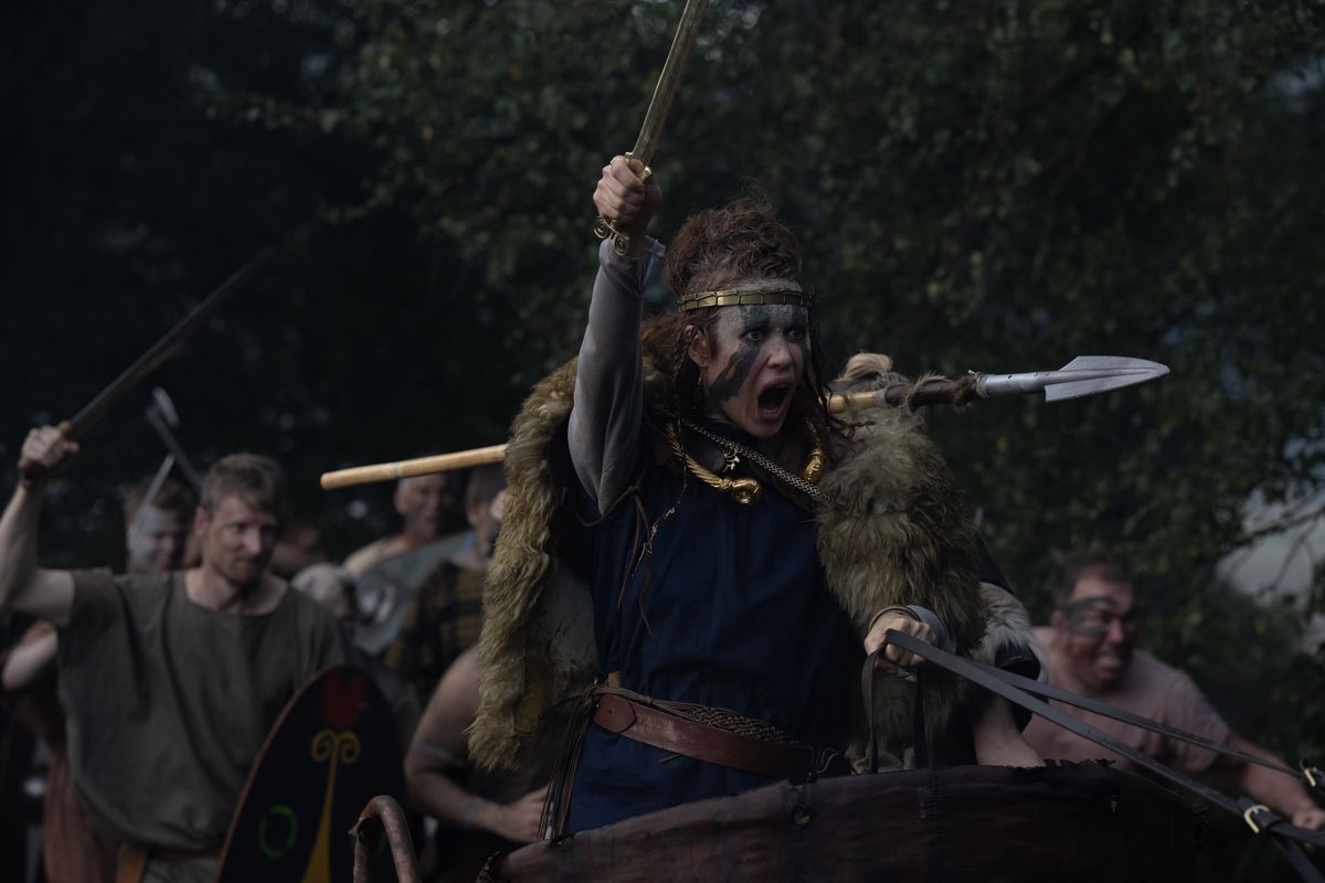 Olga Kurylenko, met blauwe schmink op, houdt een speer vast en schreeuwt in een strijdwagen terwijl krijgers achter haar staan ​​in Boudica.
