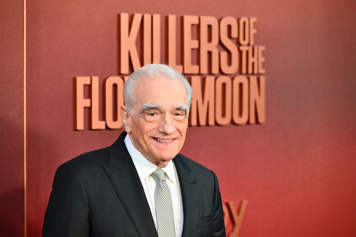 Martin Scorsese, in a black suit, smiles in front of a wall sign with the title of Killers of the Flower Moon on the red carpet at the movie’s premiere in Los Angeles