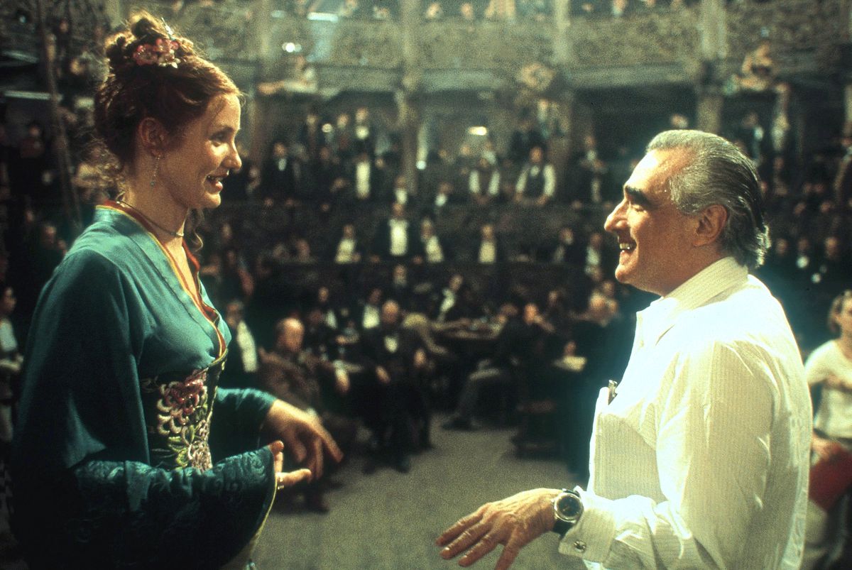 Cameron Diaz, in costume in an embroidered gown, stands opposite director Martin Scorsese on the set of The Gangs of New York, as a room full of men in dark suits, sitting in tiered wooden seating, is dimly visible in the background