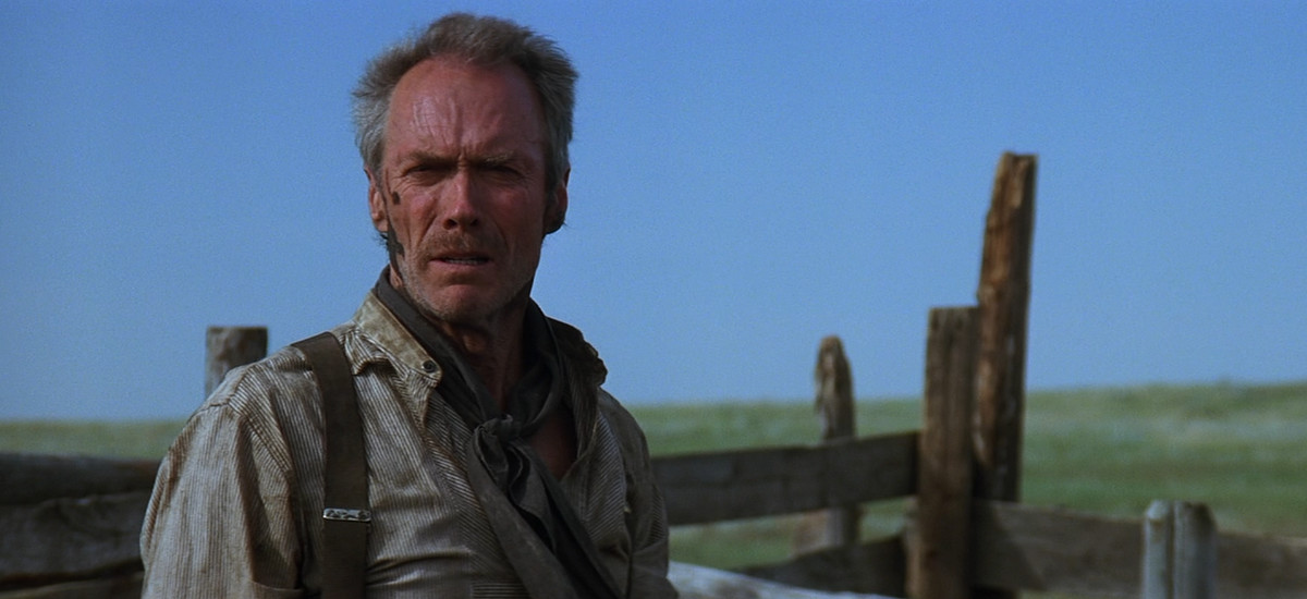 Clint Eastwood has muck on his face and looks distressed in Unforgiven, with the horizon and a large green field behind him.