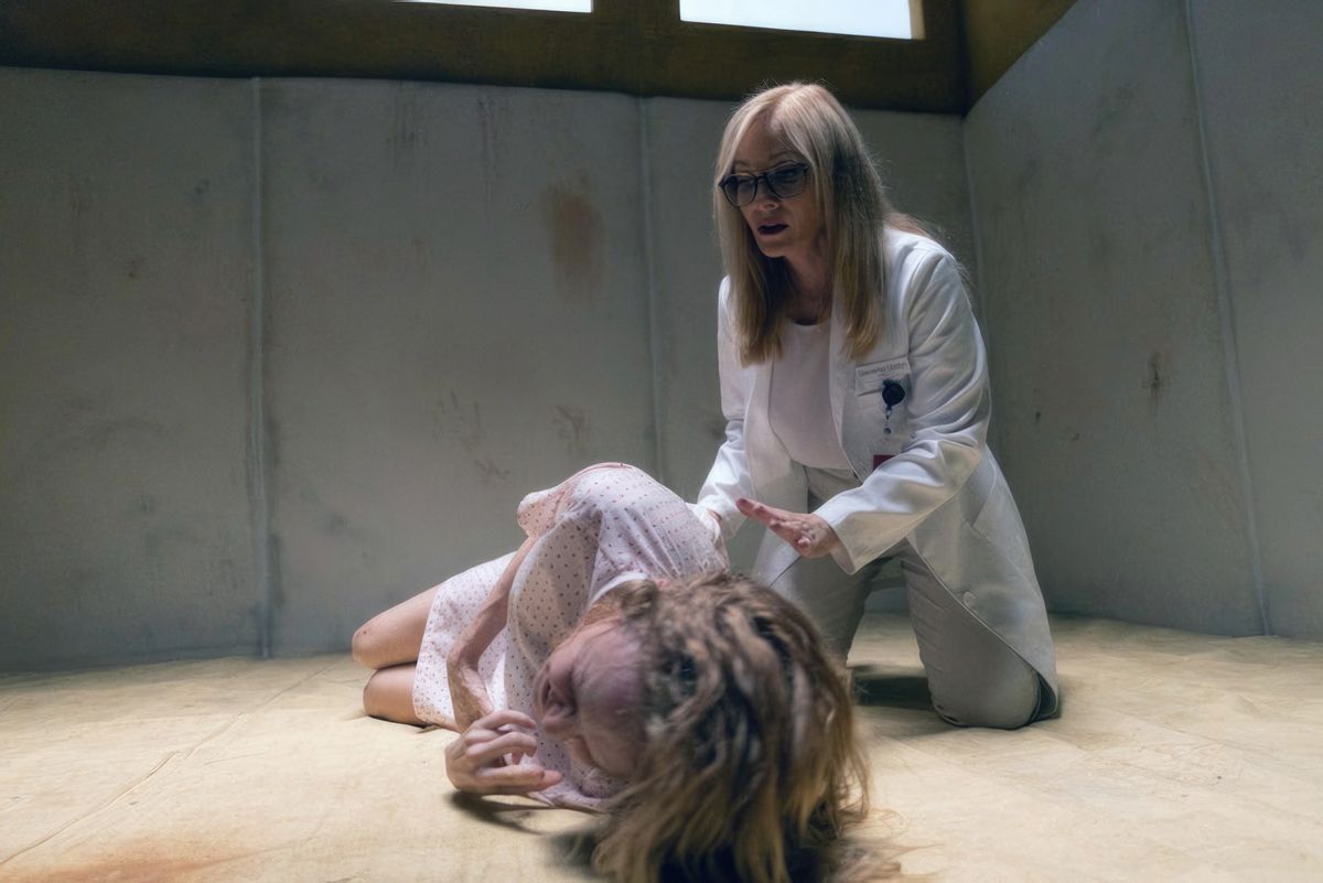 Elizabeth (Heather Graham, in a hospital gown), curls up weeping on the floor of a bare psychiatric hospital cell as a psychiatrist friend (Barbara Crampton) kneels next to her and puts out a supportive hand to her in Suitable Flesh