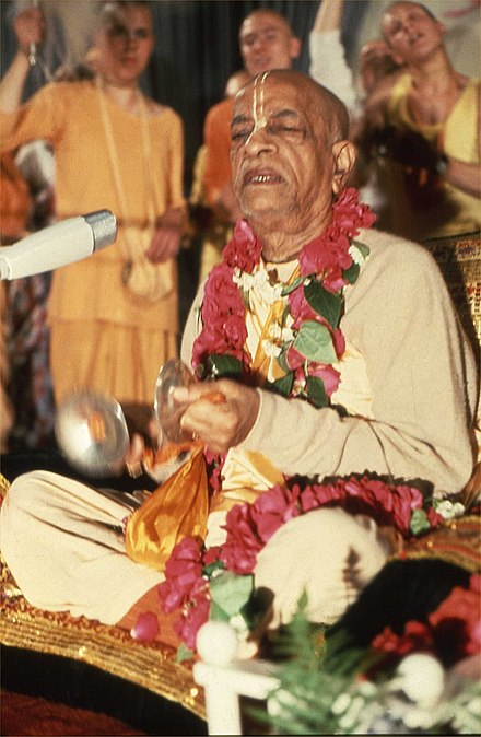Swami Prabhupada in a yellow kurta and a red garland, giving sermon. 