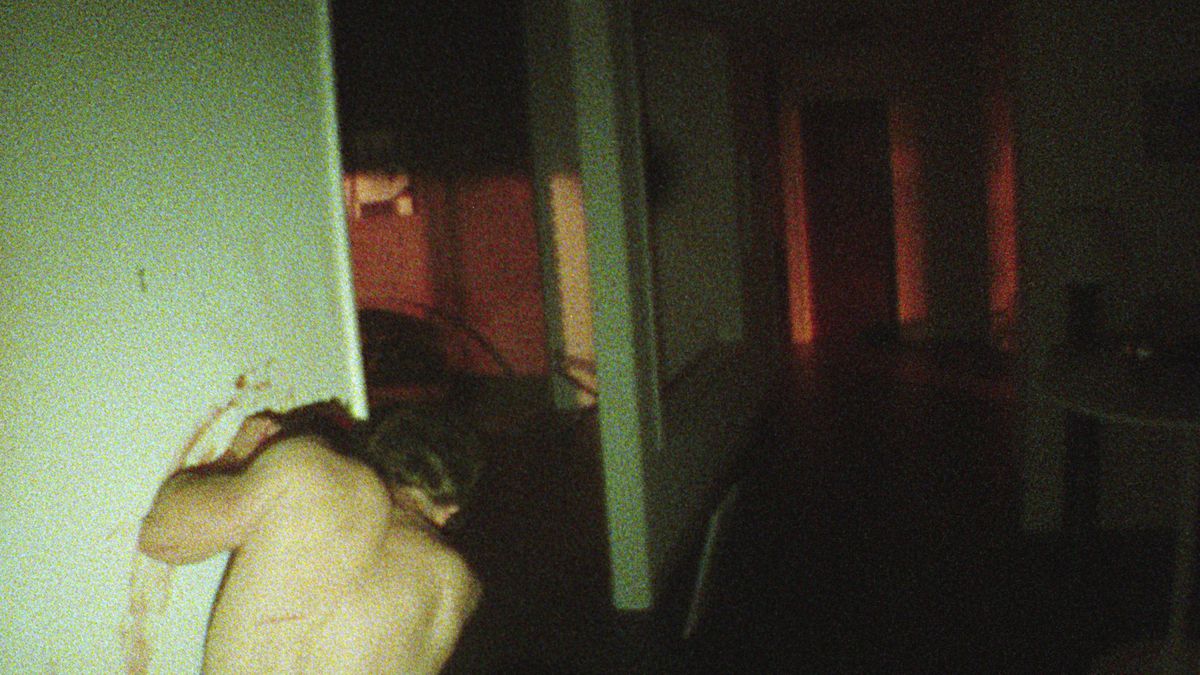 A shirtless man, seen from behind, hunches in a hallway in a dark, grainily shot apartment, leaning against a grimy greenish wall and smearing blood on it in the V/H/S/85 segment Dreamkill