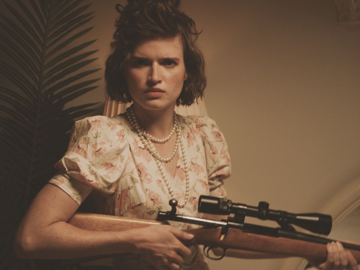 A young woman in a quaint, old-fashioned flowery dress and a long, looped pearl necklace holds a heavy-duty hunting rifle with a scope and glares into the camera in a sepia-styled portrait from the V/H/S/85 segment ‘Ambrosia’